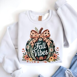 May include: White sweatshirt with a large pumpkin illustration and the text "Fall Vibes." The pumpkin is green with floral accents and a brown stem. The bow has a leopard print design. Floral accents surround the pumpkin, creating a seasonal theme.
