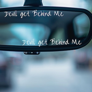 May include: A car's rearview mirror with the phrase "Devil get Behind Me" in white script. The mirror is black and reflects a blurred outdoor scene. The text is repeated on the mirror.