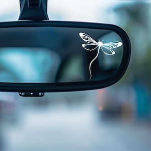 May include: A car's rearview mirror with a white dragonfly decal. The dragonfly is a simple, elegant design with delicate wings and a long, curved tail. The mirror is black and reflects a blurred outdoor scene.