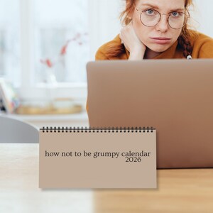 以下が含まれることがあります： 「how not to be grumpy calendar 2026」のテキストが入ったベージュ色のリング綴じカレンダーが木製の表面に置かれています。背景には、赤い髪とメガネをかけた人がノートパソコンを見ています。