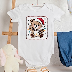 May include: White baby onesie with a Christmas-themed embroidered design. The design features a brown teddy bear wearing a Santa hat and a scarf, holding a string of lights. The onesie is hanging on a wooden rack.