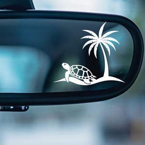 May include: White silhouette of a sea turtle swimming under a palm tree on a car's rearview mirror.