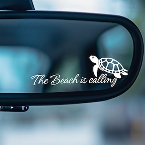 May include: A white vinyl decal featuring a sea turtle and the phrase "The Beach is calling" in a cursive font. The decal is displayed on a car's rearview mirror, against a blurred background.
