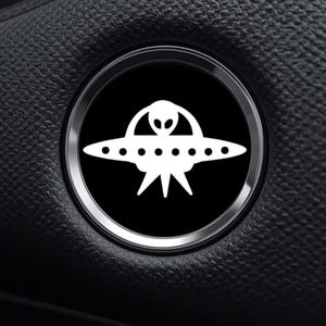 May include: A white alien spaceship design on a black background, set within a silver-colored circular frame. The image is a close-up, showing the details of the spaceship and the surrounding textured surface.