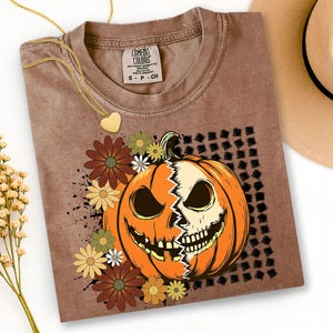 May include: Brown t-shirt with a Halloween pumpkin and skull graphic, surrounded by flowers. A gold heart pendant necklace and a hat are also visible. The t-shirt has a 'Comfort Colors' label.