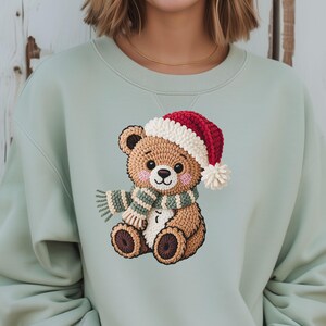 May include: A light green sweatshirt featuring a detailed embroidered teddy bear wearing a red and white Santa hat and a green and white striped scarf. The bear has a cute expression and is a festive holiday design.