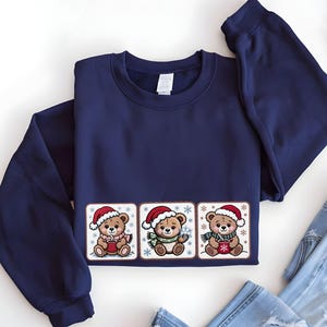May include: Navy blue sweatshirt featuring three embroidered teddy bears wearing Santa hats and scarves, holding mugs, and surrounded by snowflakes. The design is centered on the front of the long-sleeved top.
