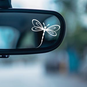 May include: White dragonfly decal applied to a car's rearview mirror. The dragonfly design features delicate wings and a slender body, creating a minimalist and elegant aesthetic. The decal is a simple, yet eye-catching car accessory.