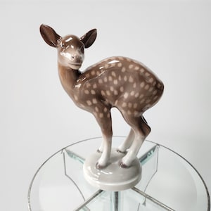 May include: A porcelain figurine of a brown and white spotted fawn standing on a glass pedestal. The fawn has a white face and large ears.