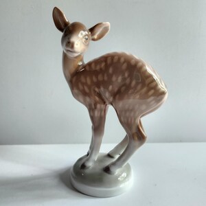 May include: A porcelain figurine of a brown and white spotted fawn standing on a white base. The fawn has a sweet expression and is looking to the side.