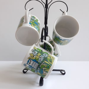 May include: Set of four white ceramic mugs with colorful floral and owl designs, displayed on a black metal mug rack.