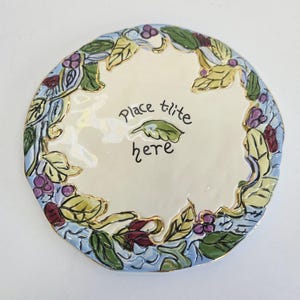 May include: Round ceramic plate with a cream-colored center and a decorative border of leaves, berries, and vines in green, yellow, blue, and purple. The plate has gold trim and the words "Place lite here" are written in black.