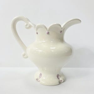 May include: A cream-colored ceramic pitcher with a curved handle and a scalloped rim. The pitcher has small purple dots around the rim and on the base. The pitcher is a decorative item.