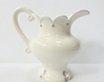 May include: A cream-colored ceramic pitcher with a curved handle and a scalloped rim. The pitcher has small purple dots around the rim and on the base. The pitcher is a decorative item.