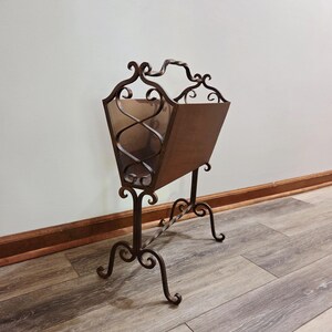 May include: A vintage magazine rack with a dark brown metal frame and a wooden container. The metal frame features ornate scrollwork and a handle at the top. The rack stands on four curved legs and is designed to hold magazines or newspapers.