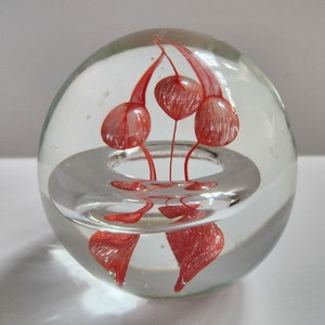 May include: A clear glass paperweight with a red and white flower design inside. The flower is suspended in the center of the paperweight.