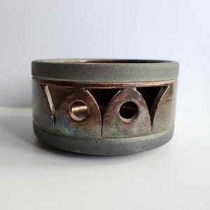 May include: A ceramic, cylindrical container with a dark gray rim and base. The body of the container is a metallic bronze color with cut-out geometric shapes. The cut-outs reveal the interior, which has a circular opening.