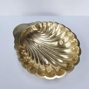 May include: A gold-colored, shell-shaped decorative bowl. The bowl has a detailed, ridged design and a scalloped edge. The interior of the bowl is smooth and reflective, and the overall shape is reminiscent of a seashell.