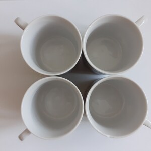 May include: Four white ceramic mugs with rounded handles. The mugs are arranged in a square formation.