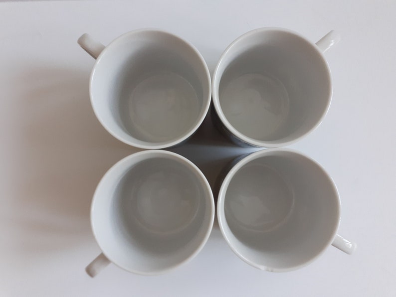May include: Four white ceramic mugs with rounded handles. The mugs are arranged in a square formation.