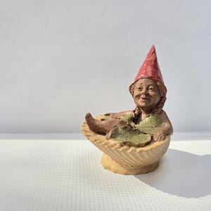 May include: A whimsical garden gnome figurine relaxing in a seashell-shaped tub. The gnome wears a red pointed hat and a green top. The figurine is made of a light brown material and has a smiling face.
