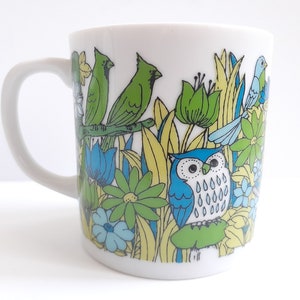 May include: White ceramic mug with a colorful design of birds, flowers, and leaves. The design features a blue owl with white spots, green birds, and yellow and blue flowers.