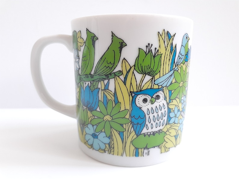 May include: White ceramic mug with a colorful design of birds, flowers, and leaves. The design features a blue owl with white spots, green birds, and yellow and blue flowers.