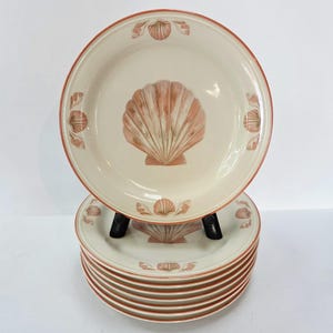 May include: A set of cream-colored ceramic plates with a seashell design. Each plate features a large coral-colored seashell in the center and smaller seashell accents around the rim. The plates have a coral-colored border.