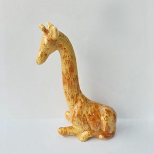May include: A ceramic giraffe figurine in a seated position. The giraffe has a long neck and is colored with shades of yellow and brown, with a speckled pattern. The figurine is resting on a white surface.