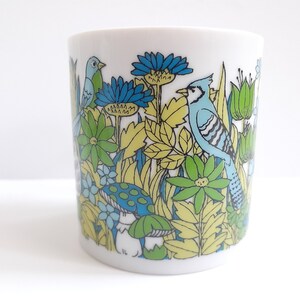 May include: A white ceramic mug with a blue, green, and yellow floral design featuring birds. The design includes a blue jay, a smaller blue bird, and green leaves and flowers.