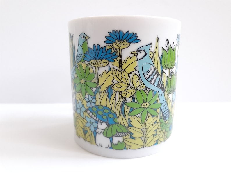 May include: A white ceramic mug with a blue, green, and yellow floral design featuring birds. The design includes a blue jay, a smaller blue bird, and green leaves and flowers.