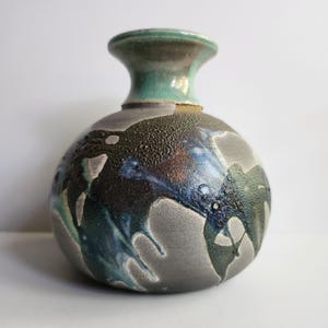 May include: A handcrafted ceramic vase with a rounded body and a narrow neck. The vase features a textured, dark gray surface with blue and green glaze accents. The rim is a muted teal color, adding a pop of color to the design.