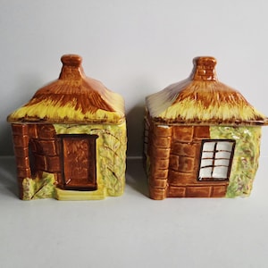 May include: Two ceramic cottage-shaped containers with thatched roofs. The containers are brown, yellow, and green, with one featuring a door and the other a window. The containers are likely for decorative or storage purposes.