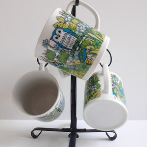 May include: Set of four white ceramic mugs with a colorful owl and floral design. The mugs are hanging on a black metal stand.