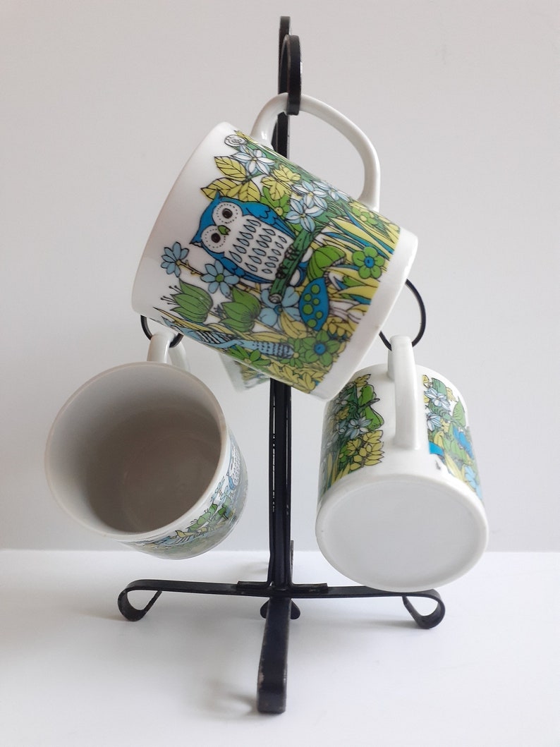 May include: Set of four white ceramic mugs with a colorful owl and floral design. The mugs are hanging on a black metal stand.