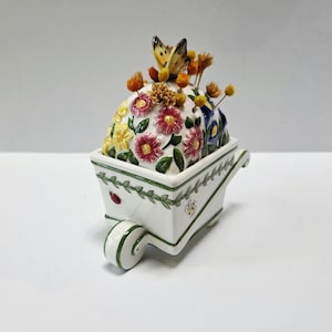 May include: A white ceramic wheelbarrow planter with a floral arrangement and a butterfly on top. The planter is decorated with green vines, a ladybug, and a bee. The floral arrangement includes red, yellow, and blue flowers.