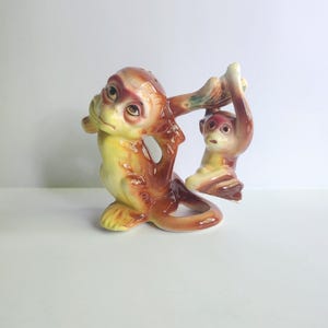 May include: A ceramic monkey figurine. One monkey is seated, with a yellow belly and brown fur, holding a branch. The second monkey hangs from the branch. The figurine has a glossy finish and is set against a white background.