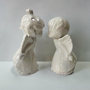 May include: Two off-white ceramic figurines of angels, facing each other in a kissing pose. Each angel has detailed wings and wears a flowing gown. The figurines are on a plain white background.