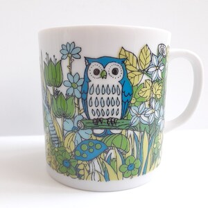 May include: A white ceramic mug with a blue and green owl design. The owl is surrounded by flowers, leaves, and mushrooms.