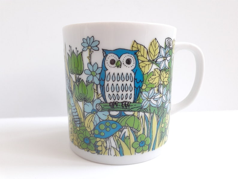 May include: A white ceramic mug with a blue and green owl design. The owl is surrounded by flowers, leaves, and mushrooms.