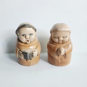 May include: Two ceramic monk figurines. One monk is reading a book labeled "BIBLE" and the other is in a prayer position. Both are wearing brown robes and have light skin tones. The figurines are on a white background.