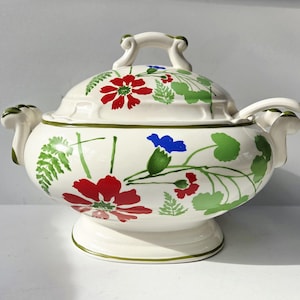 May include: A white ceramic soup tureen with a lid, adorned with hand-painted red, blue, and green floral designs. The tureen has two handles and a pedestal base, perfect for serving soups and stews.
