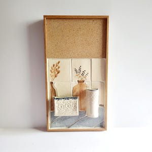 May include: A framed wall art piece featuring a corkboard top and a ceramic tile section below. The tile section depicts vases with floral designs, a decorative soap dish, and a cylindrical container. The frame is made of wood.