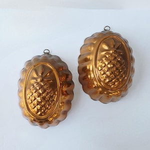 May include: Two antique copper pineapple molds. Each mold has an oval shape with a scalloped edge and a small ring for hanging. The pineapple design is embossed in the center of each mold, showcasing the fruit's texture and shape.