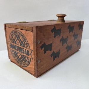 Può includere: Una scatola di shortbread in legno con finitura marrone scuro. La scatola presenta sagome nere di cani Scottie e le parole "All Natural Ingredients Shortbread" stampate sul davanti. Ha un pomello di legno sulla parte superiore.