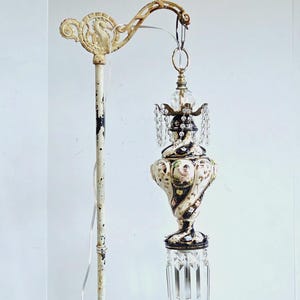 May include: An antique floor lamp with a cream-colored pole and a decorative, ornate top. The lamp features a hanging, decorative glass and crystal fixture with black and white accents and a floral design. The lamp's metal arm has a gold finish.