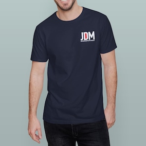 May include: Navy blue t-shirt with the white and red text "JDM JAPANESE DOMESTIC MARKET" printed on the upper left chest. The shirt is a solid color and has short sleeves.