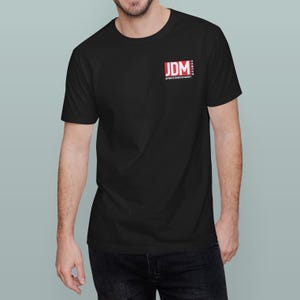 May include: Black t-shirt with a small red and white "JDM" logo on the upper left chest. The logo includes the text "Japanese Domestic Market". The shirt is short-sleeved and has a crew neck.