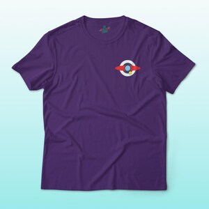 Puede incluir: Una camiseta morada con un peque&ntilde;o gr&aacute;fico de un dise&ntilde;o rojo, blanco y azul. El gr&aacute;fico es una representaci&oacute;n estilizada de un planeta o cuerpo celeste. La camiseta tiene mangas cortas y cuello redondo.