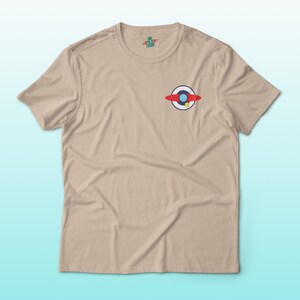 Puede incluir: Una camiseta de color beige con un peque&ntilde;o gr&aacute;fico colorido en el pecho. El gr&aacute;fico representa un dise&ntilde;o rojo, blanco y azul con un acento amarillo. La camiseta tiene mangas cortas y cuello redondo.